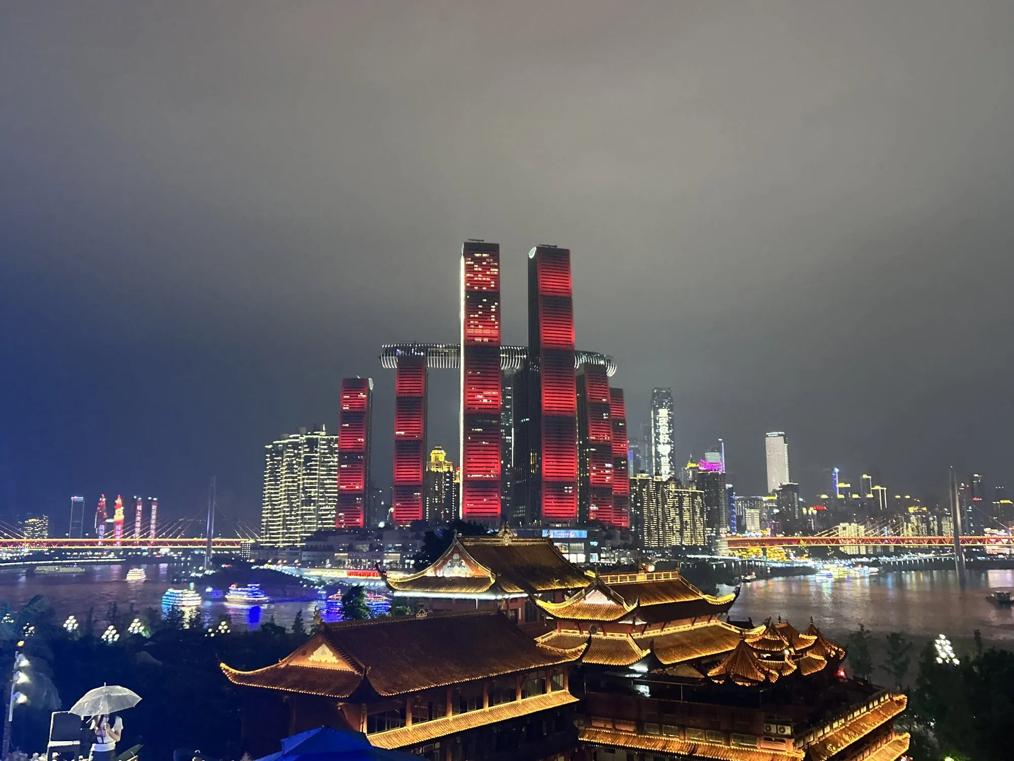 A city skyline at night with red-lit towers above glowing traditional rooftops by a river.