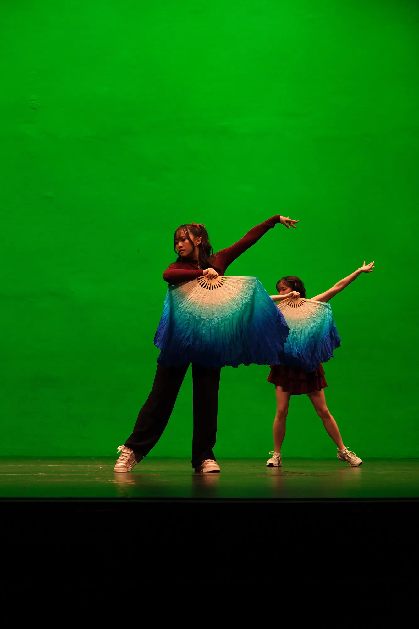 Two dancers on stage against a green backdrop, each holding a blue and white silk fan.