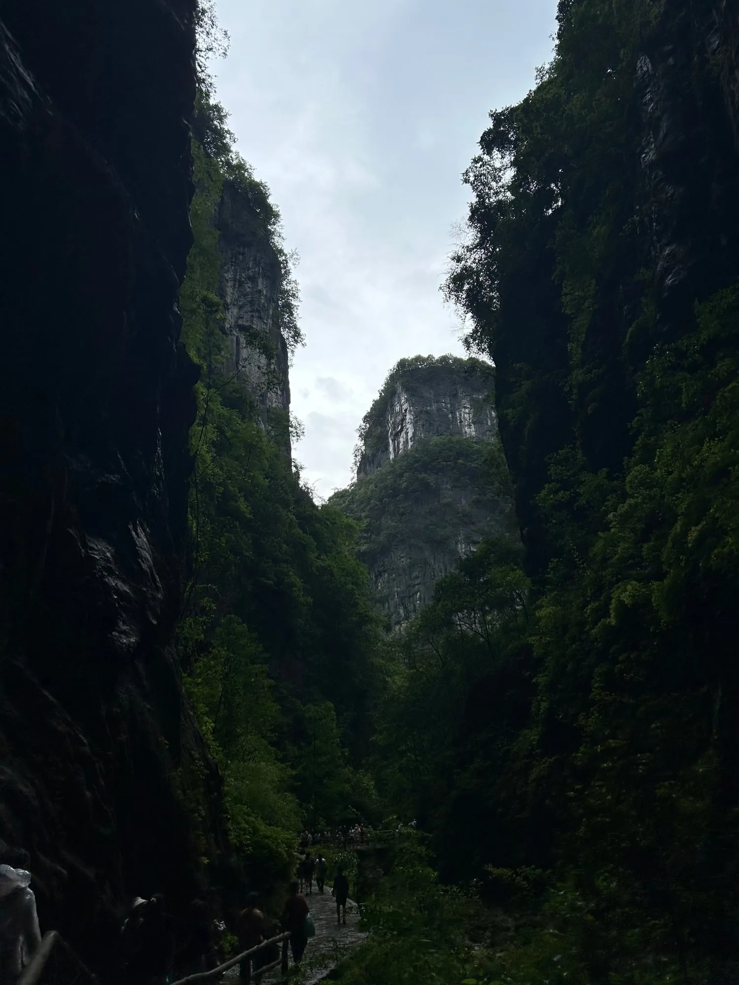 Hikers on a path at the bottom of a narrow gorge, green cliffs rising on both sides toward a pale sky.