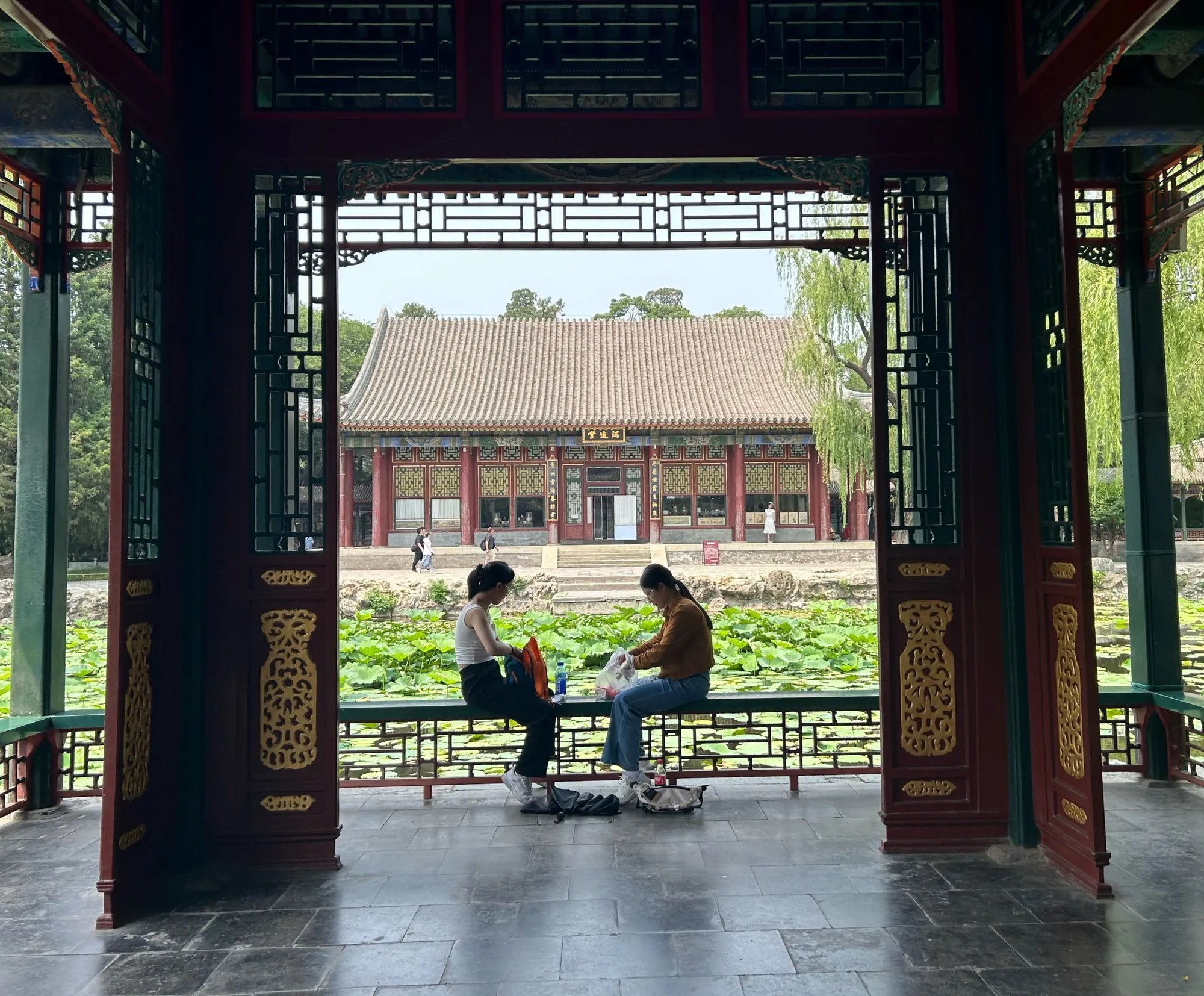 Two people sitting in a painted wooden pavilion, a lotus pond and a traditional hall beyond.
