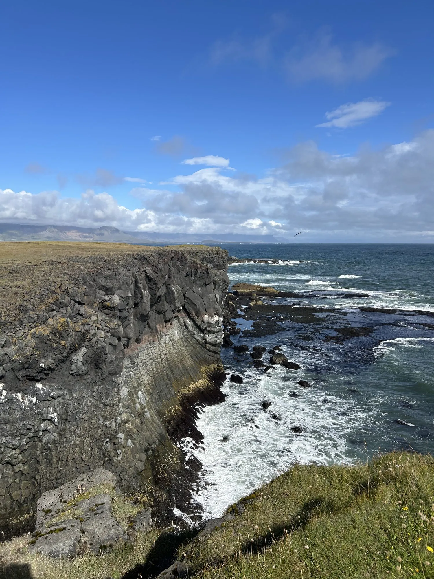 Dark rocky cliffs meeting white surf under a partly cloudy sky.