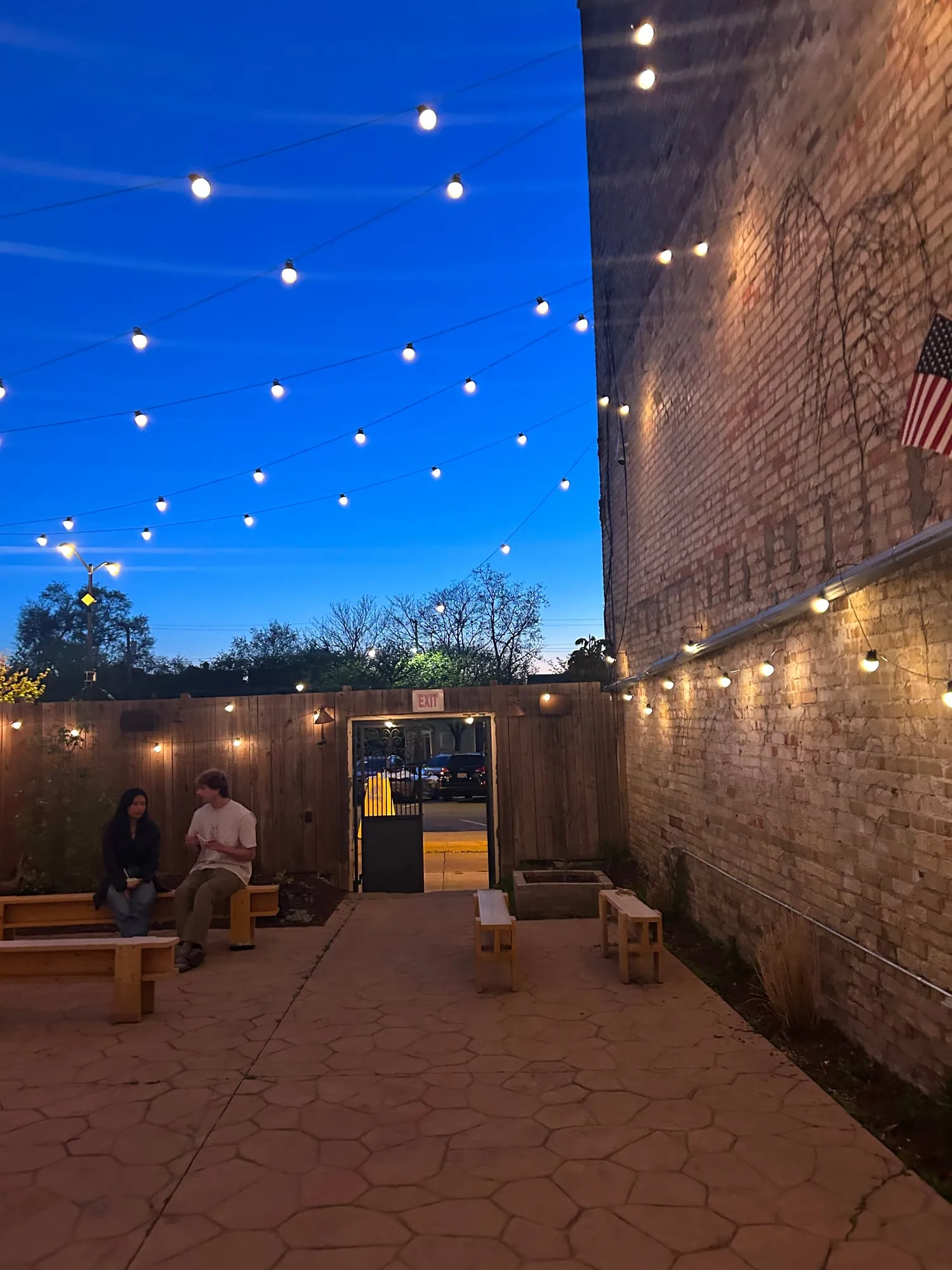 A brick-walled patio at dusk under rows of glowing string lights, two people talking on a bench.